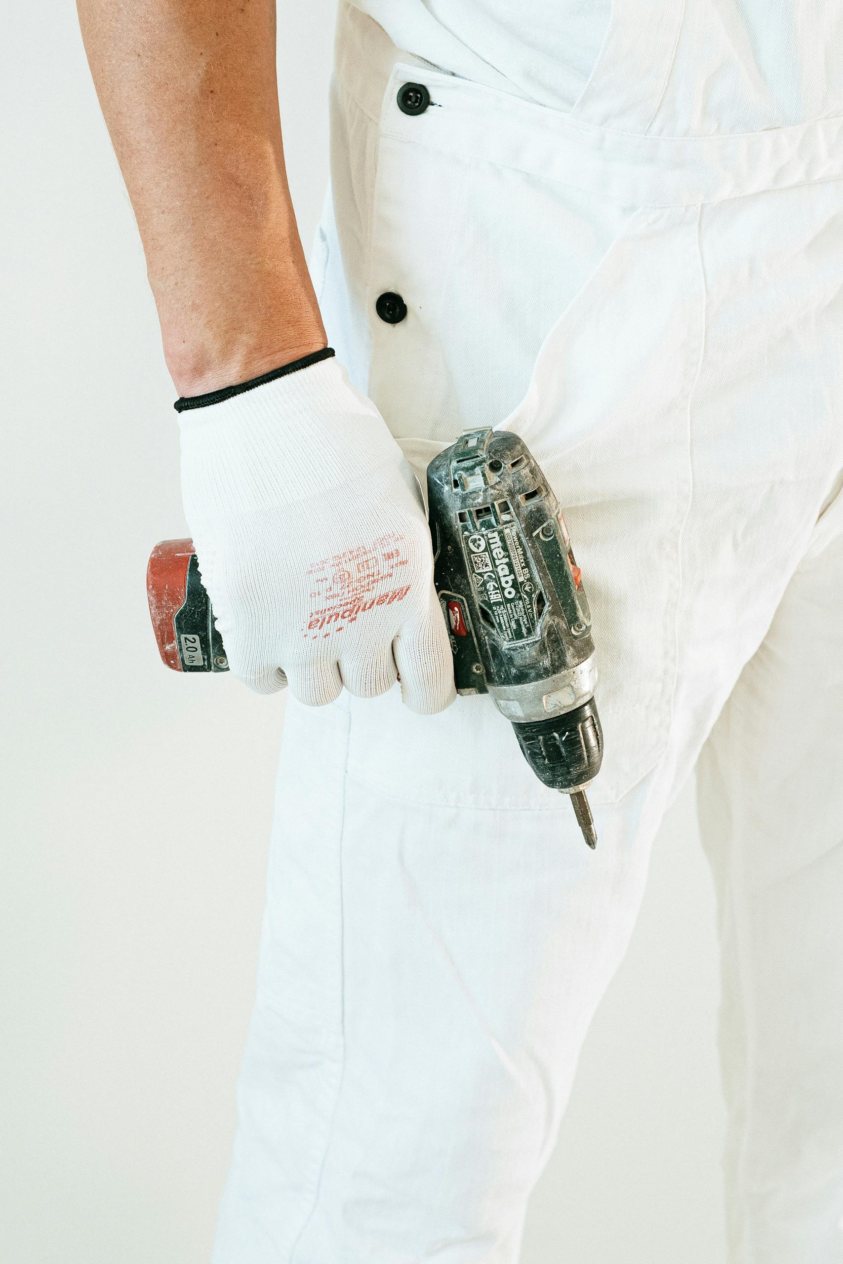 Worker's hand holding power drill, wearing white overalls and gloves. Close-up view.